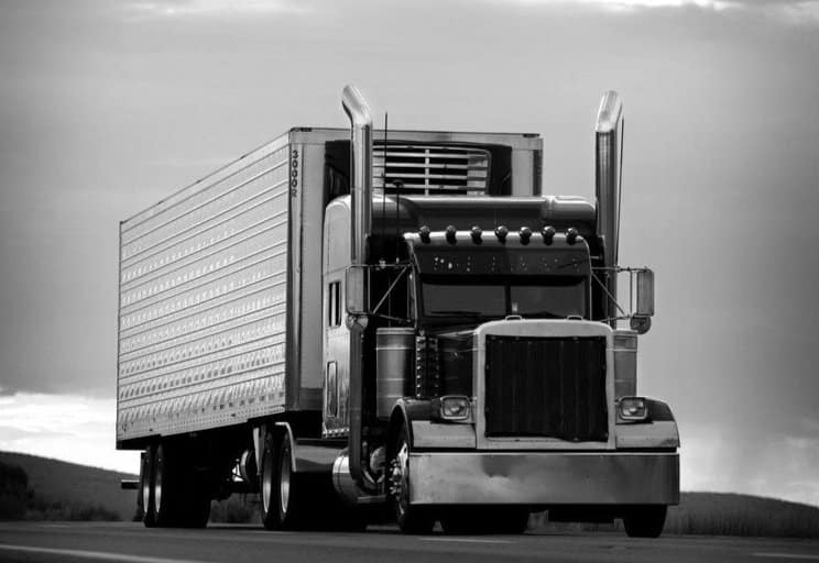 A black and white image of a large semi-truck with a trailer driving on an empty road. The truck has tall exhaust stacks on both sides of the cab and a prominent grille at the front. The cloudy sky in the background creates a dramatic atmosphere.