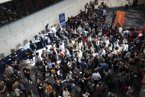 A large group of people gather at a bustling conference hall with various informational booths and tables. Attendees engage in conversations and network, creating a lively atmosphere. Banners and signs are visible in the background.