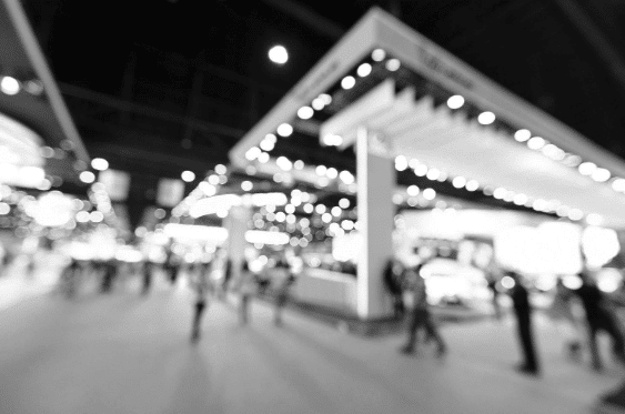 A blurred black-and-white image of a busy indoor convention or exhibition center. Numerous people are walking past lighted booths and displays, with vibrant lights and signs creating an energetic atmosphere.