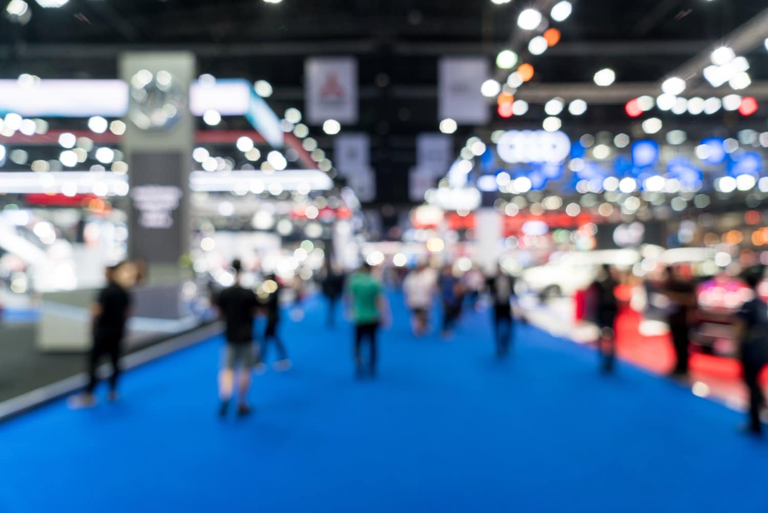 A blurred image of a busy exhibition hall with people walking on a blue carpet. Various booths and bright lights are visible in the background, creating a vibrant, bustling atmosphere.
