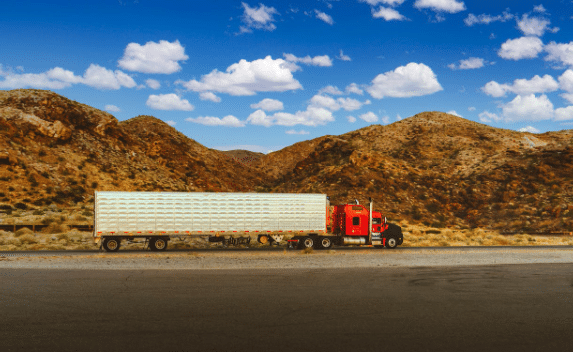 A red semi-truck with a long white trailer is parked on a road in a desert landscape. The background features rugged, brown hills under a bright blue sky with scattered white clouds.