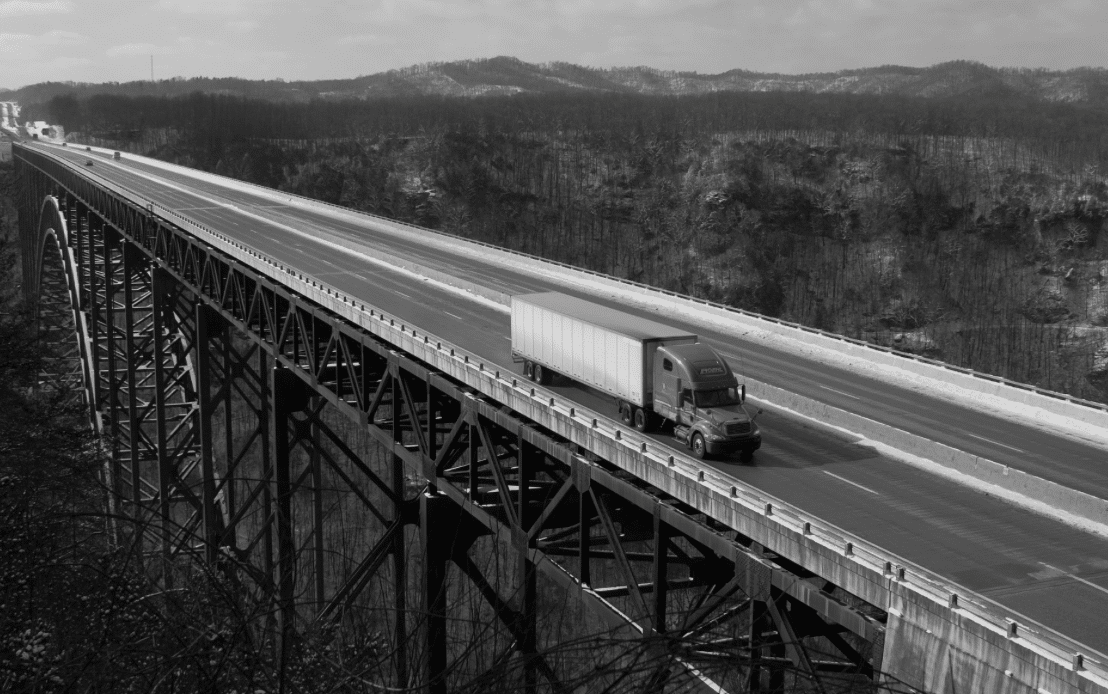 A semi-truck drives across a tall, expansive bridge spanning a deep valley. The bridge has a metal lattice structure beneath the roadway. In the background, forested hills extend towards the horizon under a partly cloudy sky.