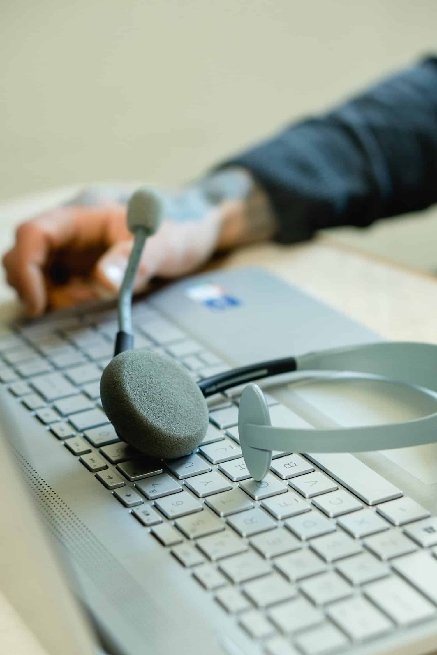 A headset with a microphone rests on a laptop keyboard. A person's hand, tattooed and wearing a gray sleeve, is typing on the keyboard. The scene suggests a work-from-home or remote communication setup.