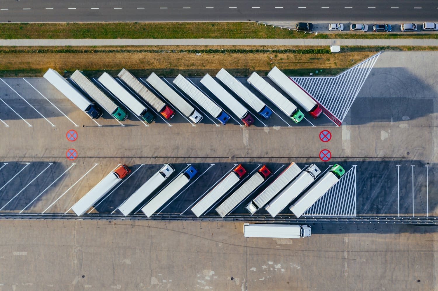 Aerial view of several semi-trucks parked diagonally in marked bays at a truck stop. The trucks are of different colors, and a single semi-truck is driving through the lot. Nearby, a road runs parallel with parked cars along its side.