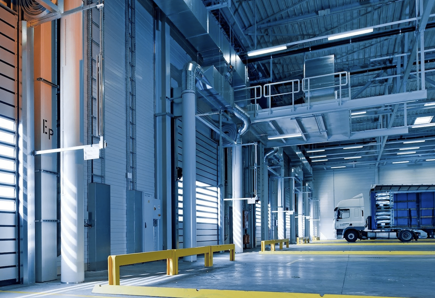 A modern, spacious warehouse with high ceilings, steel beams, and large roll-up doors. A white truck with a blue cargo container is parked inside, next to yellow safety barriers. Fluorescent lights and sunlight through windows illuminate the space.