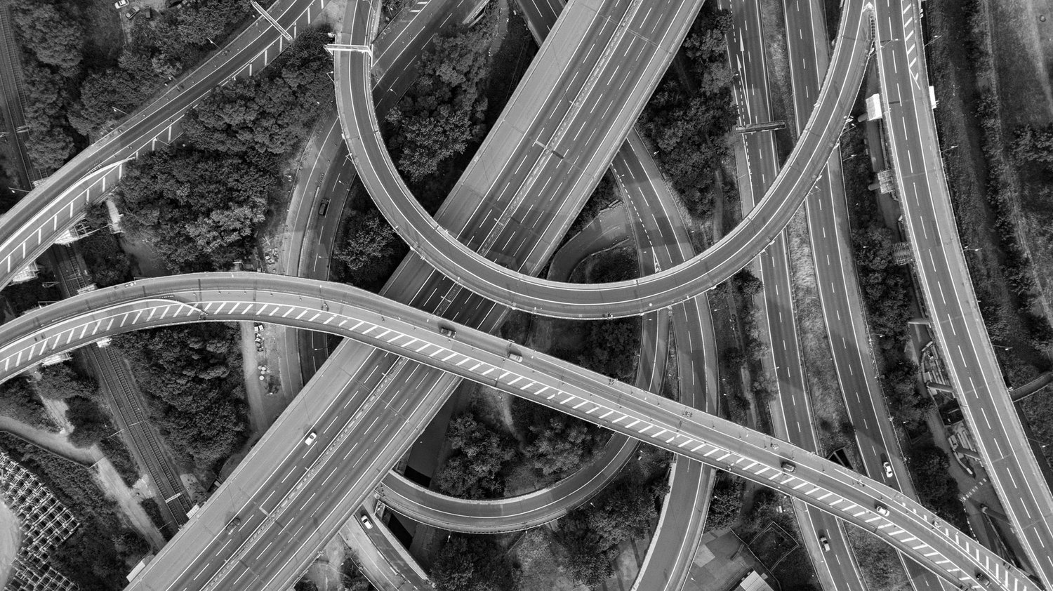 Aerial view of an intricate, multi-level highway interchange. The image shows several roads and ramps intertwining, curving, and crossing over one another, surrounded by patches of trees and greenery. The traffic appears light, with a few vehicles on the roads.