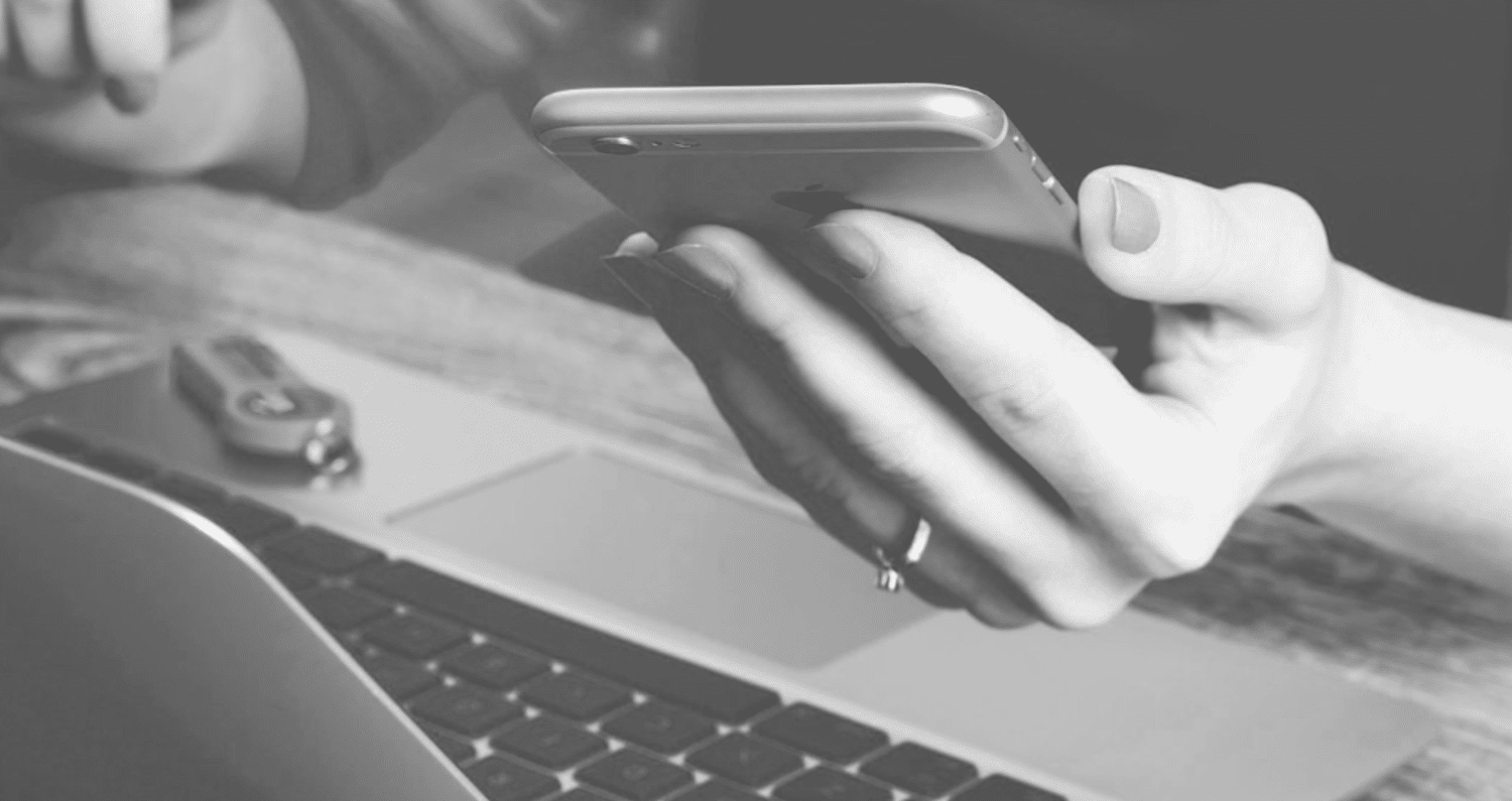 A grayscale image showing a close-up of a person holding a smartphone in one hand and a laptop partially visible below. The focus is on the hand holding the smartphone, with a USB flash drive placed on the laptop keyboard in the background.