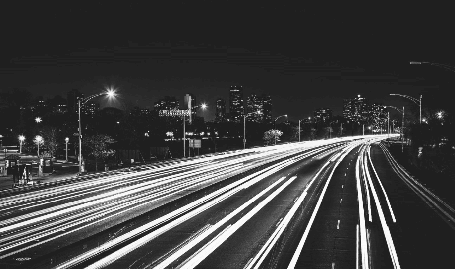 A black and white photo capturing light trails on a busy highway at night. Headlights from cars create bright, continuous lines of light, contrasting with the dark surroundings. The city skyline with illuminated buildings is visible in the background.